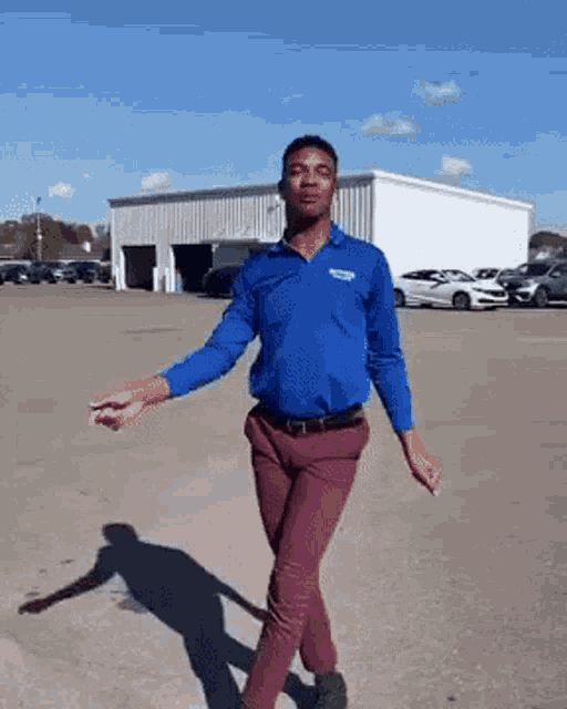 a man in a blue shirt is dancing in a parking lot with cars parked in the background