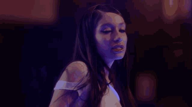 a woman in a white top is standing in a dark room with purple lights behind her .