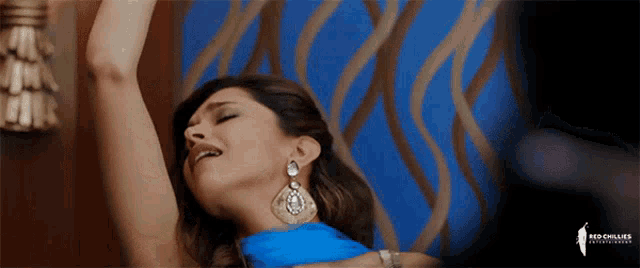 a woman in a blue dress is dancing in front of a blue wall with the word red chillies on the bottom