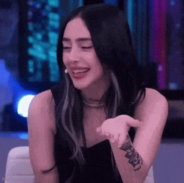 a woman with long black hair and a tattoo on her arm is sitting on a chair and smiling .