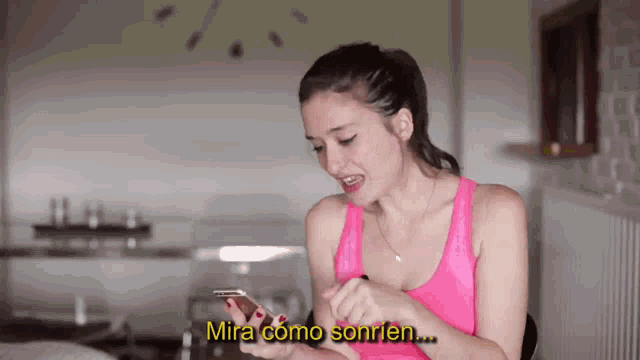 a woman in a pink tank top is smiling while looking at her cell phone