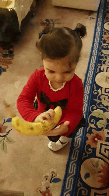 a little girl in a red shirt holds a banana in her hand