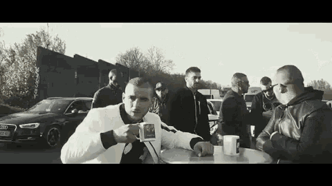 a man is sitting at a table with a cup of coffee while a group of men are standing around him .