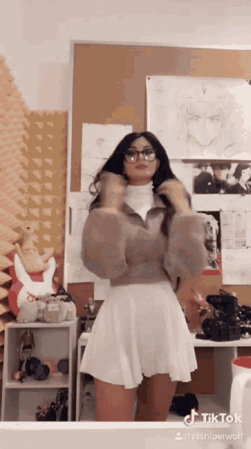 a woman wearing glasses and a white skirt is standing in front of a wall with pictures on it .