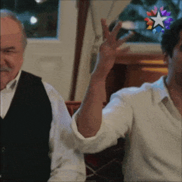 two men are sitting at a table and one is raising his hand in the air