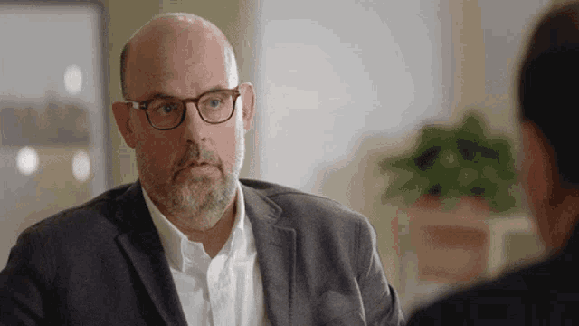 a bald man wearing glasses and a suit talks to another man