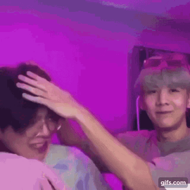 two men are standing next to each other in a room with purple lights . one of the men is touching the head of the other .
