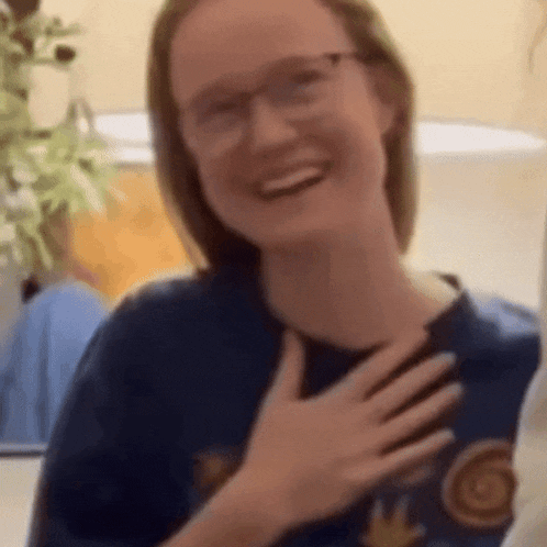 a young woman wearing glasses is smiling and holding her hand to her chest .