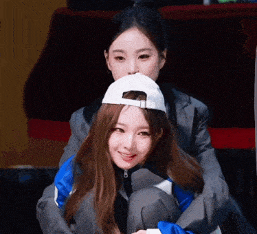 two girls are sitting next to each other with one wearing a baseball cap .
