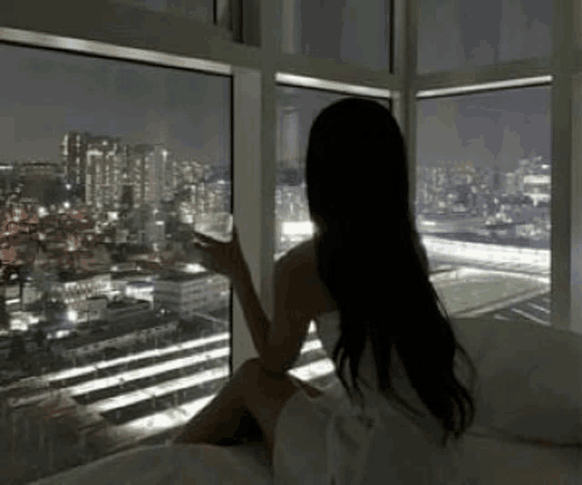 a woman is sitting on a bed with a glass of wine looking out a window at night .