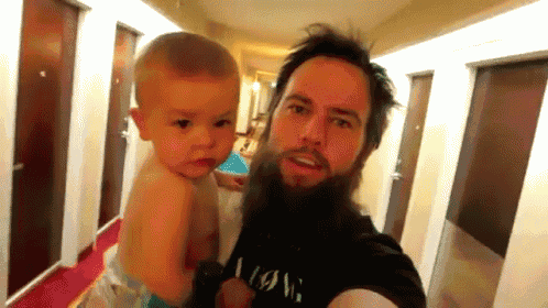 a man with a beard is holding a baby in a hallway wearing a black shirt that says wow