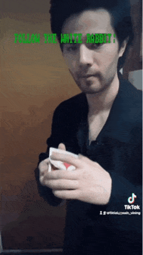 a man in a black jacket is holding a playing card in his hand and says follow the white rabbit .