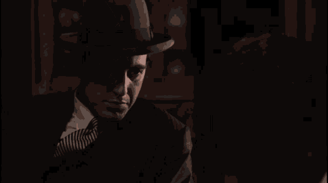 a man in a suit and hat is talking to another man in a dark room