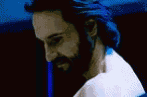 a man with a beard and blue hair is standing in a dark room and looking at the camera .
