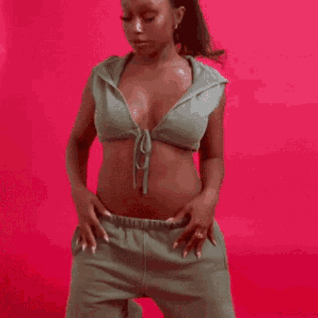 a woman in a crop top and sweatpants is laying on her stomach in front of a pink background .