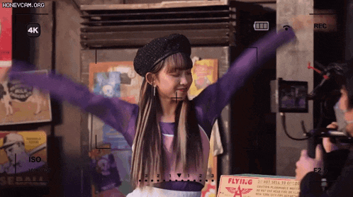 a woman in a purple shirt is standing in front of a camera with her arms outstretched ..