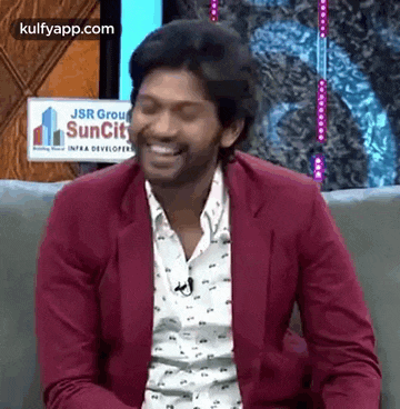 a man in a red jacket and white shirt is sitting on a couch laughing .