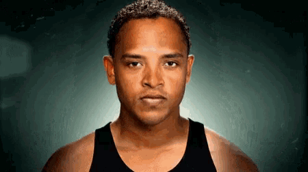 a man in a black tank top looks at the camera with a serious look on his face