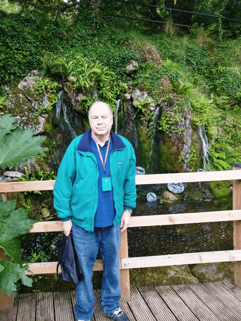 a man in a green jacket with a lanyard around his neck poses in front of a waterfall