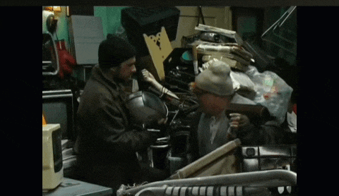 two men are standing in a room with a pile of junk including a microwave