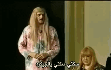 a man with long blonde hair is standing next to a woman with blonde hair and arabic writing on the screen .