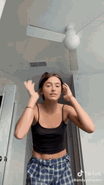 a woman in a black tank top and plaid pants is standing in a room with a ceiling fan .