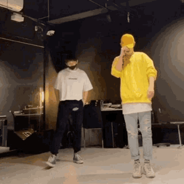 two men are dancing together in a dark room . one of the men is wearing a yellow sweatshirt and a yellow hat .