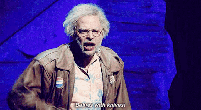 a man with gray hair and glasses is standing on a stage and says babies with knives !