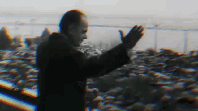 a man in a black coat is standing in front of a crowd of people and waving his hands .