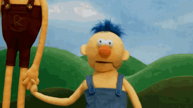 a yellow puppet with blue hair is holding the hand of another puppet with a letter r on his shorts
