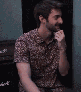 a man in a floral shirt is sitting in front of a marshall amplifier and talking into a microphone .