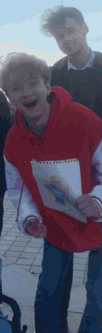 a young man in a red hoodie is holding a drawing and smiling while standing next to another young man .