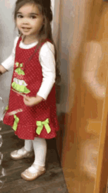 a little girl in a red dress with green bows is standing next to a door