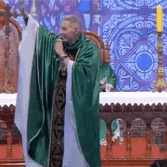 a priest in a green robe is holding a microphone in front of a stained glass wall .