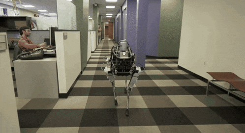 a man sits at a desk in an office cubicle while a robot walks down a hallway