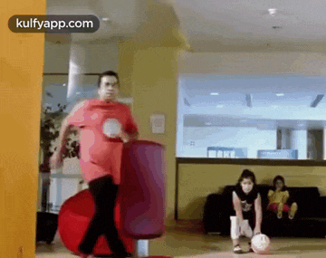 a man in a red shirt is kicking a soccer ball while a woman watches .