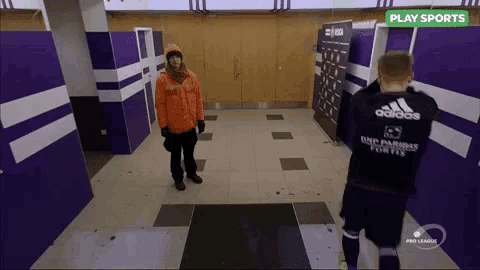 a man in an orange jacket stands next to a man in an adidas jersey in a locker room