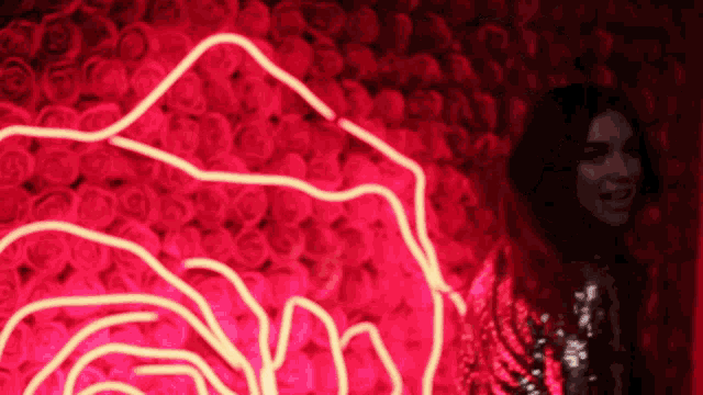 a woman stands in front of a wall of red roses with a neon rose in the background
