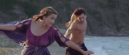 a woman in a purple dress is running with a shirtless man in the water
