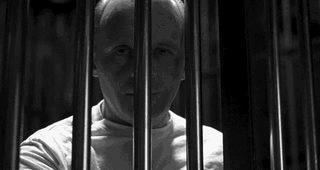 a man in a white shirt is behind bars in a prison cell .