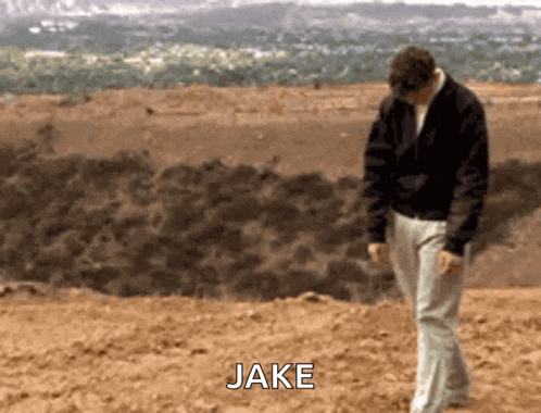 a man in a black jacket is walking in the dirt with the name jake written on the ground behind him .