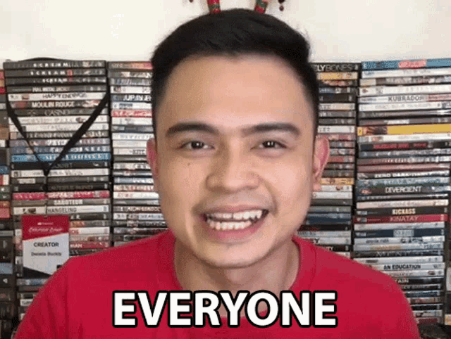 a man in a red shirt is smiling in front of a stack of dvds and says everyone .