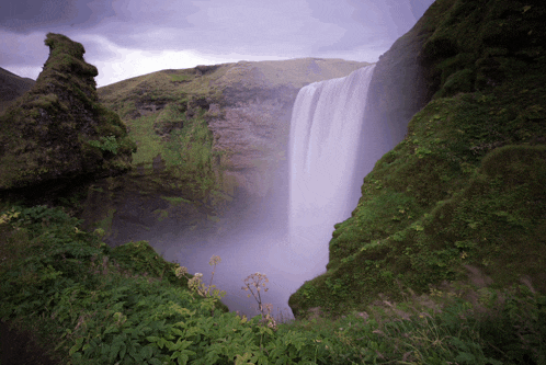 a waterfall is surrounded by lush greenery and rocks on a cloudy day