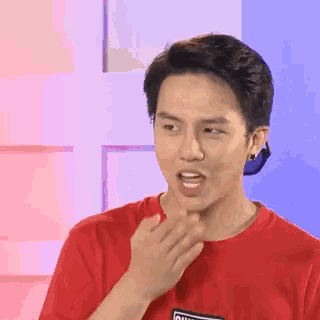 a young man in a red t-shirt is making a funny face and covering his mouth with his hand .