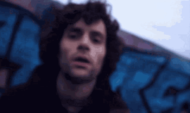 a man with curly hair stands in front of a graffiti wall with the letter r on it