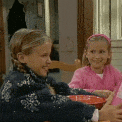 two little girls are sitting at a table with bowls of food and smiling