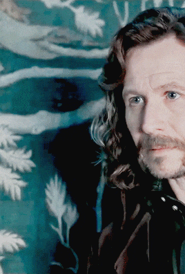 a man with long hair and a beard looks at the camera in front of a painting
