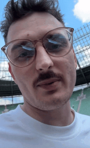 a man wearing glasses and a white shirt is standing in front of a stadium