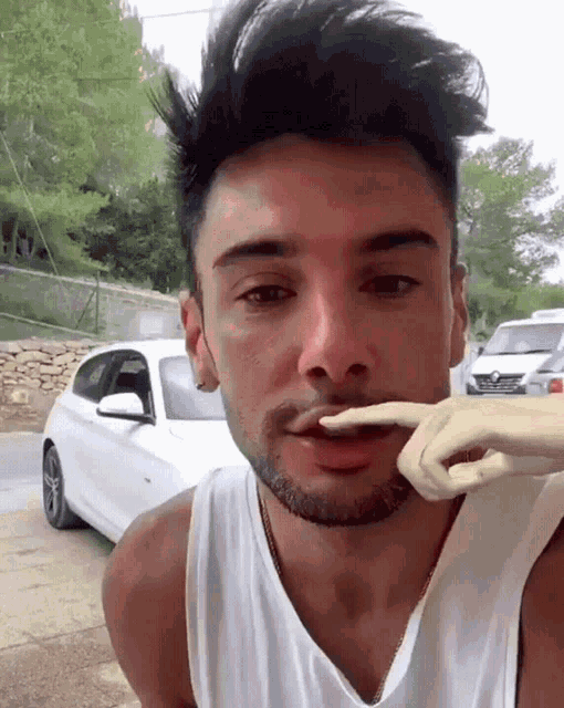 a man with a beard and a white tank top has a white car parked behind him