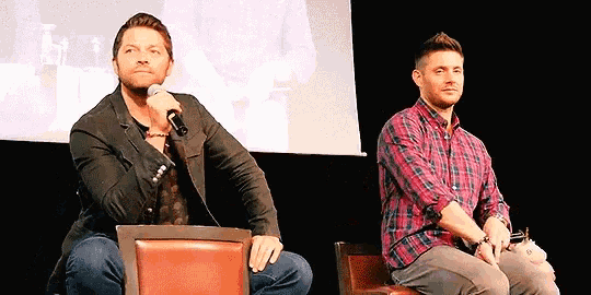 two men are sitting next to each other on a stage and one of them is holding a microphone .